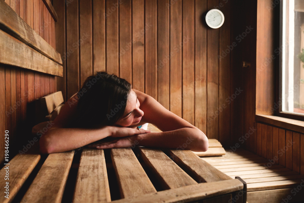 A beautiful woman wearing a white towel takes a sauna: The sauna is made of wood with a large window with a view of the snow. Concept of: relax, vacation, wellness center.